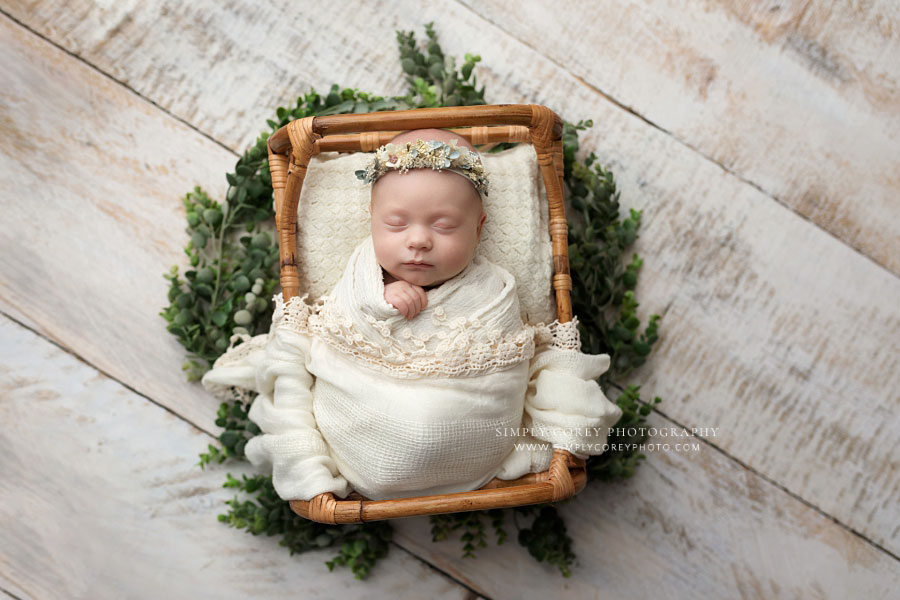 newborn photographer near Carrollton, GA; baby girl in white with rattan bed and greenery