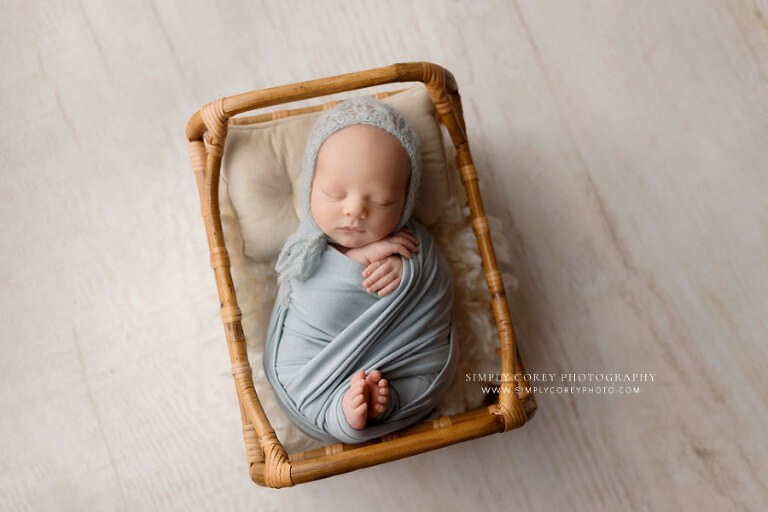 Powder Springs newborn photographer, baby boy in blue swaddle and hat with rattan bed