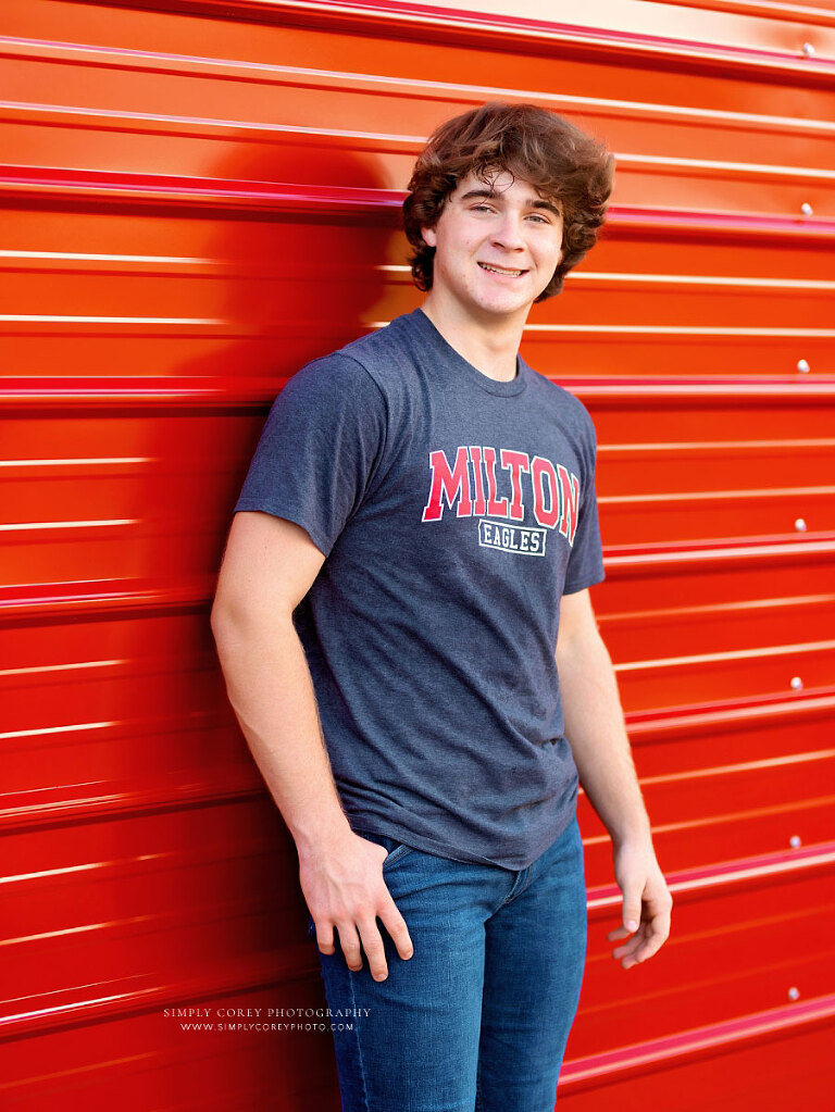 senior portrait photographer near Atlanta, teen in Milton High shirt near red metal wall